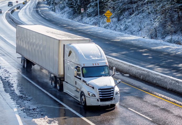 Big rig long haul gray semi truck tractor transporting commercial cargo in dry van semi trailer going on the wet slippery road with water from melting snow and winter snowy trees on the hills