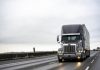 Big rig stylish industrial dark gray semi truck with turned on headlights transporting cargo in dry van semi trailer running on the twilight wet road with light reflection surface in rain weather
