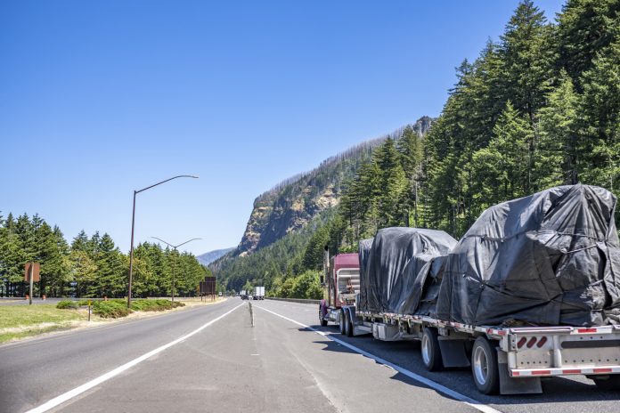 Industrial burgundy classic big rig semi truck tractor with extended cab for truck driver rest transporting fastened cargo on step down semi trailer driving on green highway road in Columbia Gorge
