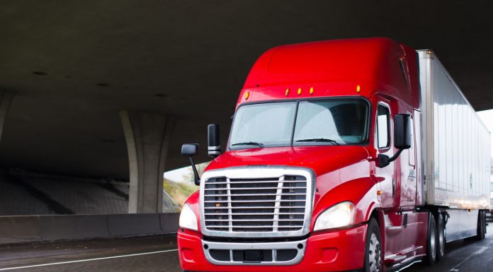 A modern big rig semi truck for long haulage with a high cabin for improving aerodynamic characteristics moves under the bridge across a multi-lane highway transporting a dry van semi trailer with commercial cargo to the place of delivery