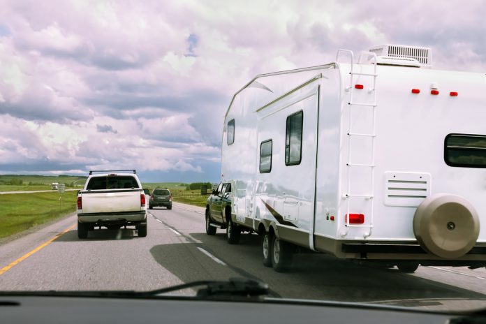 RV being pulled by truck and other vehicles on road with cloudy skies on sunny day.